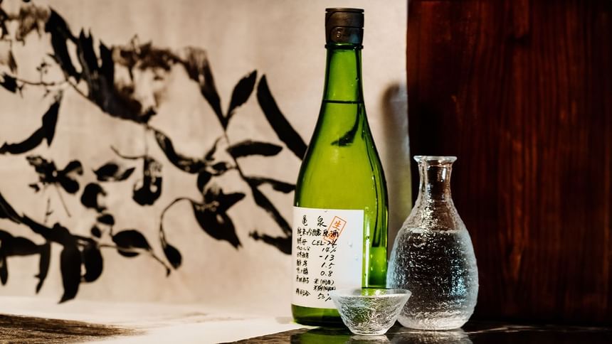 A green sake bottle and a clear glass carafe near a textured black-and-white art piece at The Kitchens