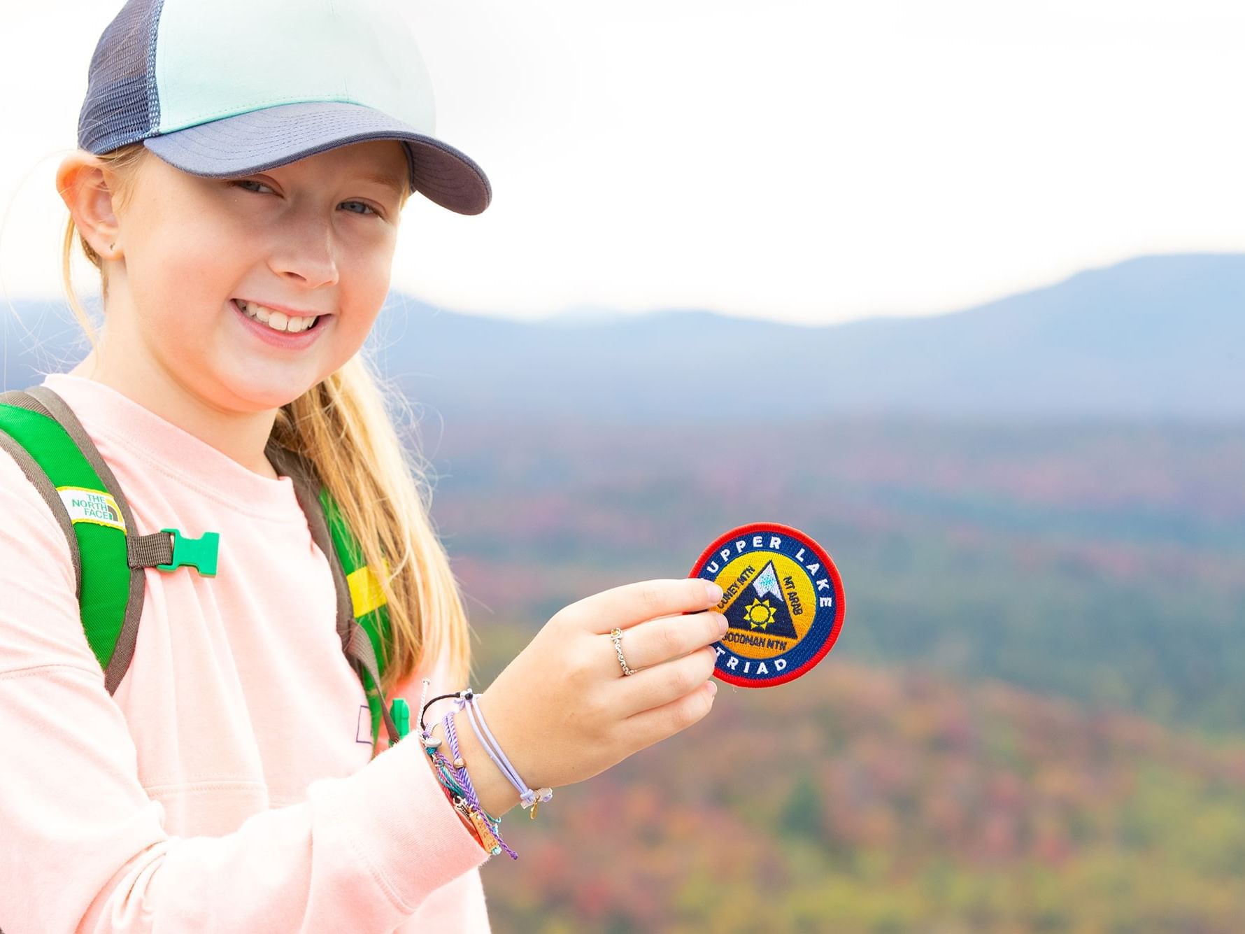 Girl with a hat and a patch that says Tupper Lake Triad, smiling in front of a mountain range near High Peaks Resort
