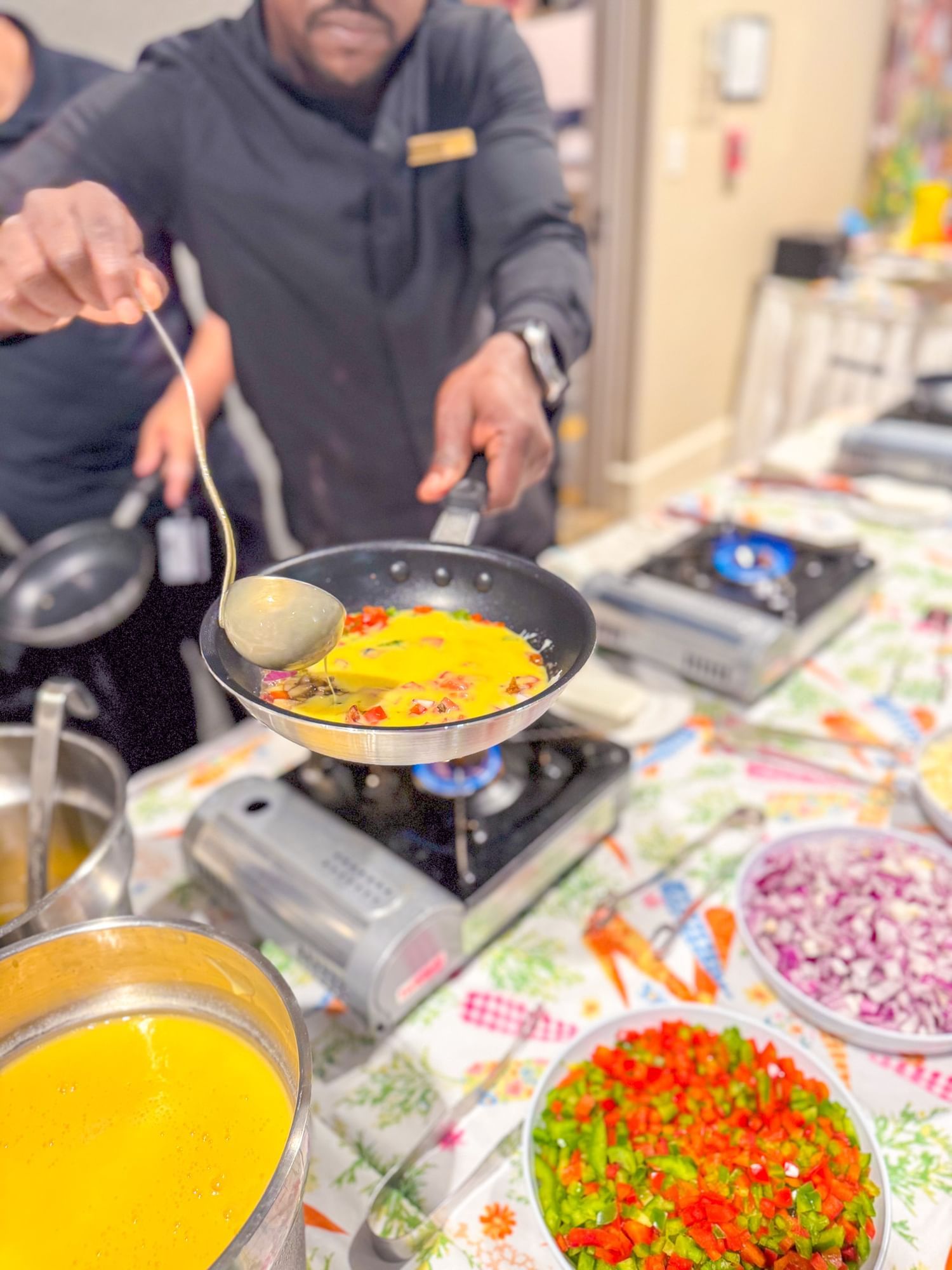 A chef in a black chef’s jacket ladles eggs into a pan over a table with burners and bowls of vegetables.  