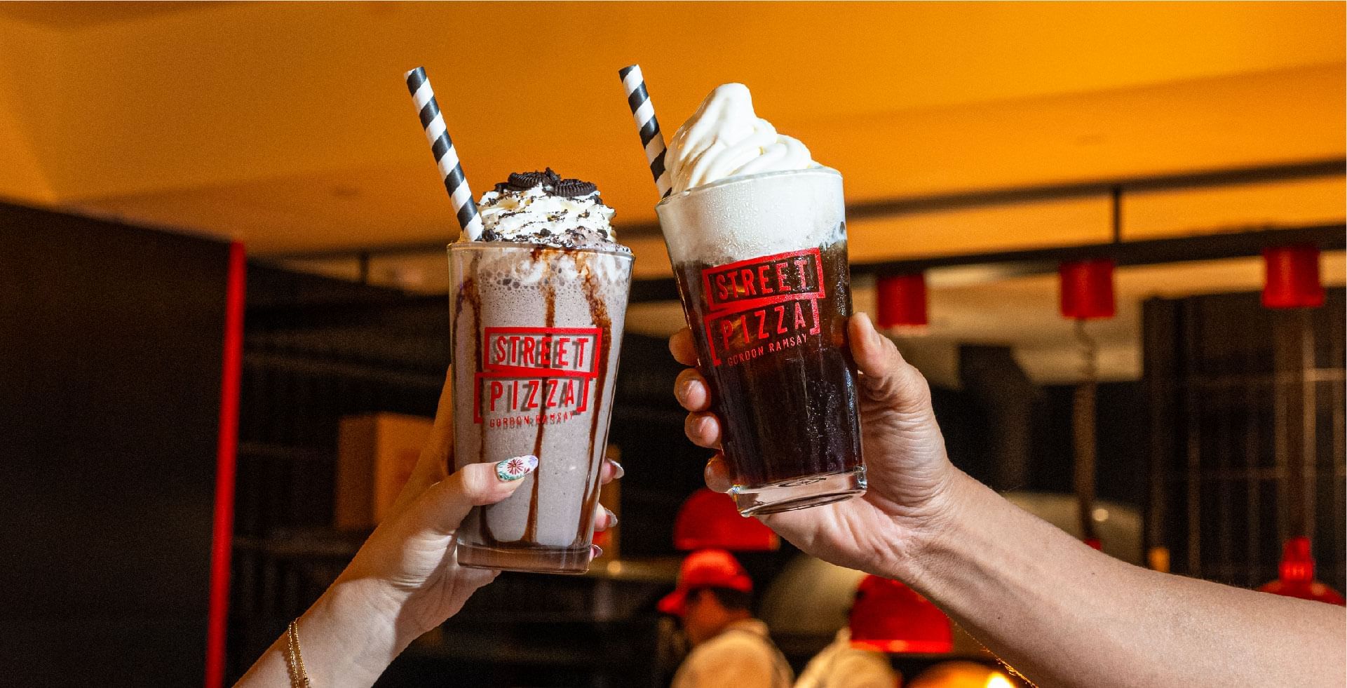 Two hands holding creamy milkshakes topped with whipped cream in Gordon Ramsay Street Pizza at Sunway Resort