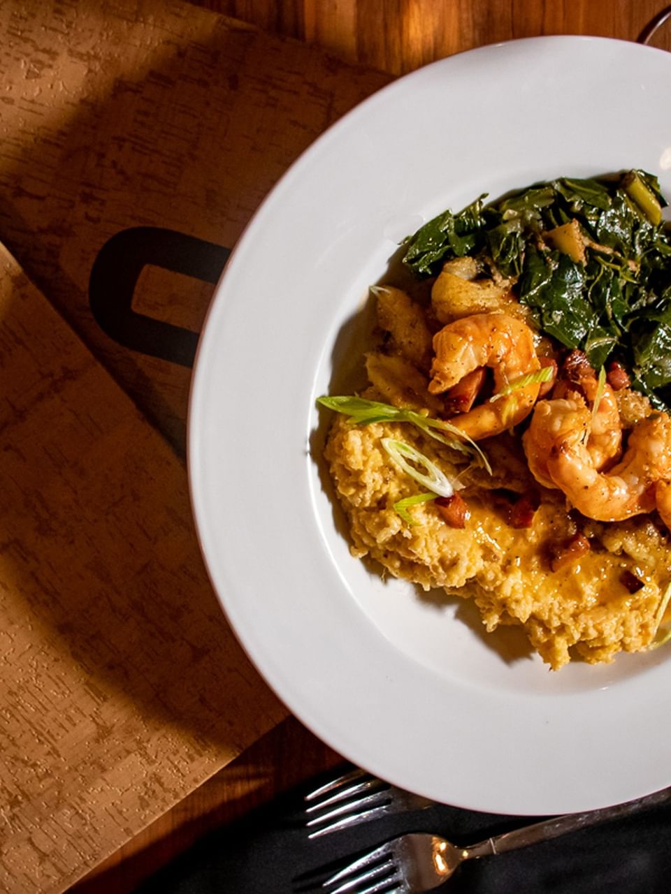 A plate of shrimp and grits served with greens on a wooden table in Cora's Restaurant at The White House Hotel