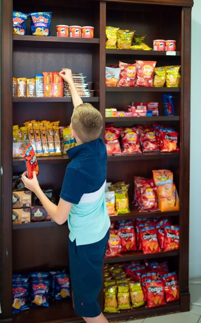 Boy picking snacks from a large wooden pantry shelf filled with potato chips and treats at Rodd Moncton