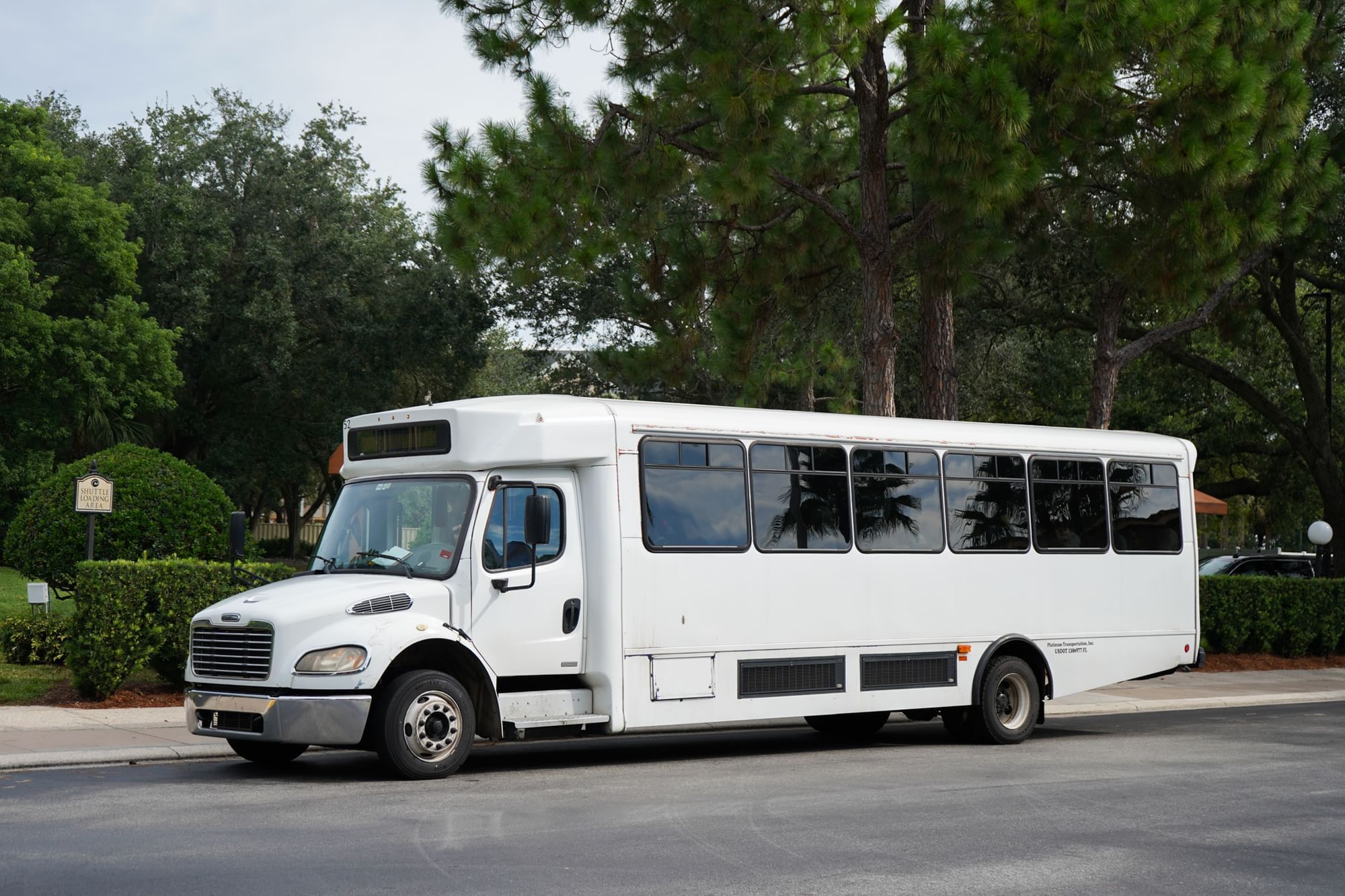 A  white shuttle bus is parked on a paved area with trees and greenery in the background. Staying at Rosen Inn Lake Buena Vista offers the advantage of complementary scheduled shuttle service to Orlando theme parks. 