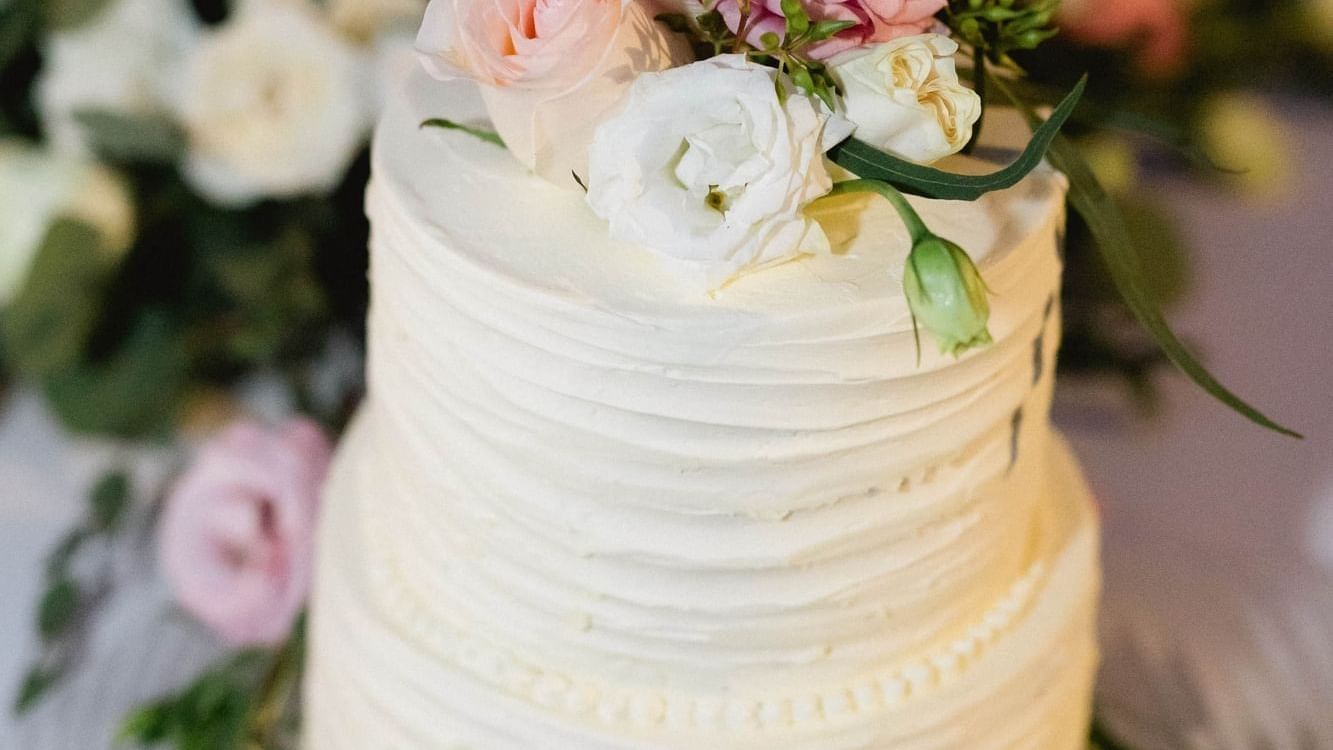 Two-tiered white cake with textured frosting, decorated with pink and white roses and greenery at The Explorean Resorts