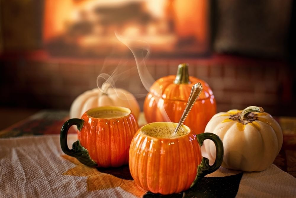 Two pumpkin-shaped mugs with steaming hot liquid alongside three pumpkin-shaped decor items and a fireplace in the background.