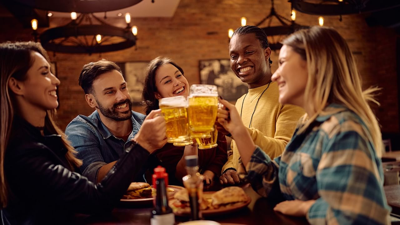 Five friends toast with beers at a restaurant table with food and condiments.