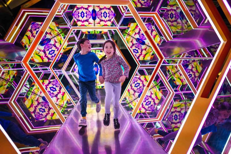 Two children jumping joyfully in a colorful illuminated mirror maze with hexagonal patterns at Warwick Le Crystal Montreal