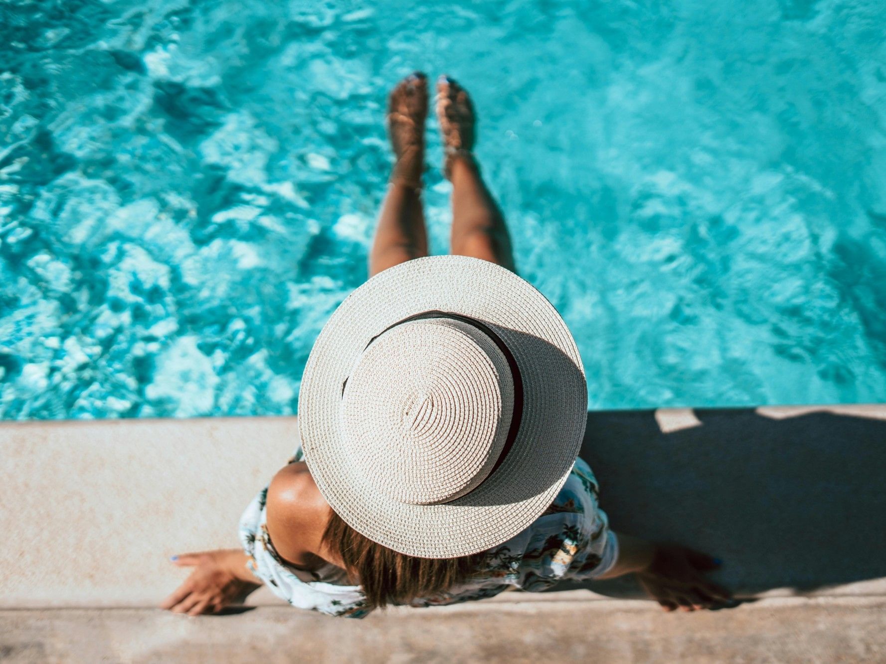 Overhead view of a woman wearing a white sun hat and floral shirt sitting on the edge of a bright blue swimming pool with her legs in the water at Hotel Grand Chancellor Townsville.