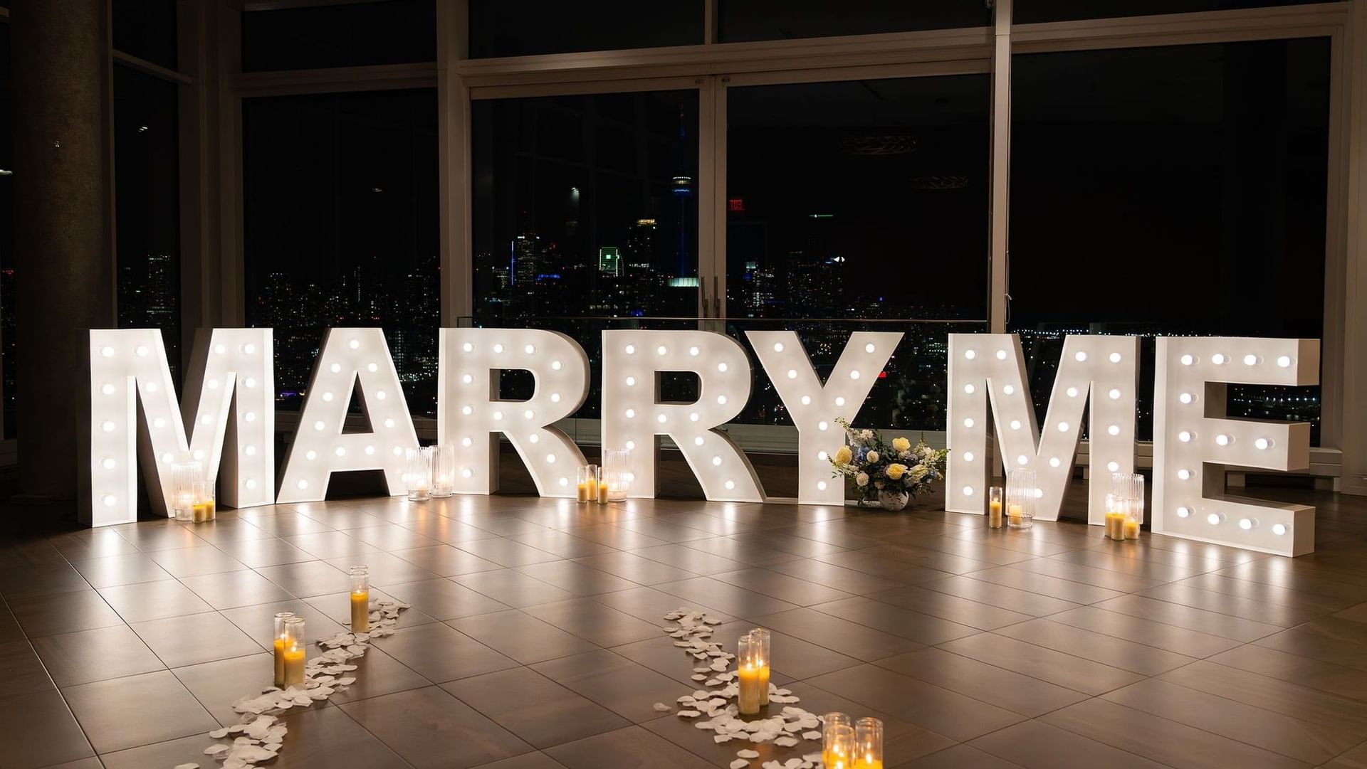 Large illuminated MARRY ME letters with candles and flower arrangement against nighttime city skyline at Hotel X Toronto