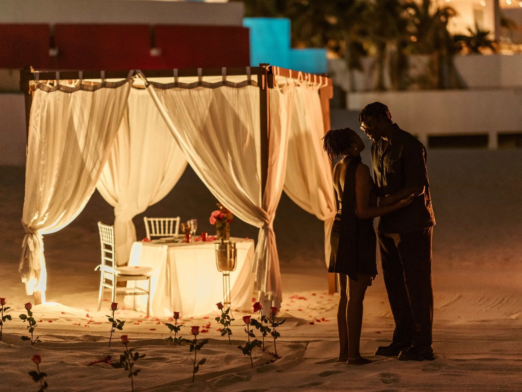 A romantic candlelit dinner setup on the beach at Marquis Los Cabos Resort with a couple embracing by a draped cabana