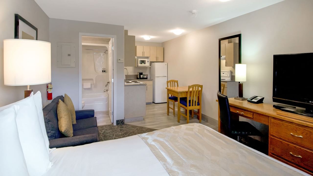 A room featuring a queen-sized bed, a small sofa, a kitchenette and a bathroom, a functional layout with ample natural light.