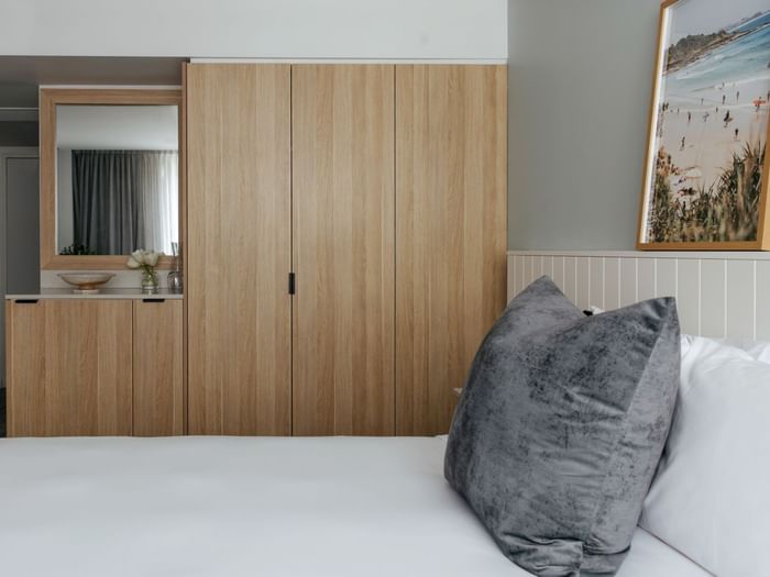 Village Queen at Elysium Manly by a mirrored dresser under framed beach art beside a grey velvet pillow on the bed