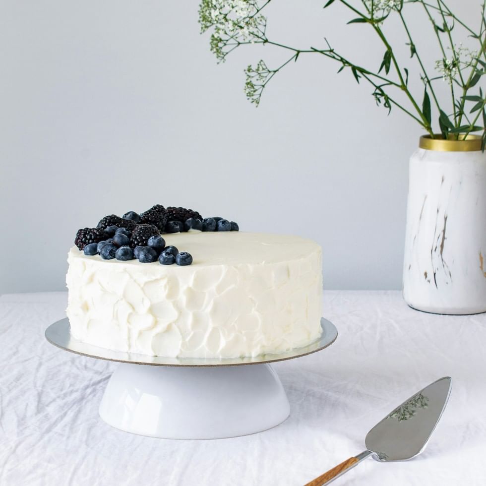 White cake with berries on a cake stand next to a vase with flowers for Mother’s Day Baking Party.