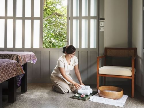 Spa room at The Surin Phuket with a practitioner preparing towels and a wooden bowl, set in a natural environment