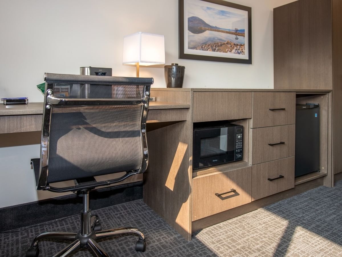 Hotel room with desk, chair, lamp, microwave, fridge, and picture frame on wall.
