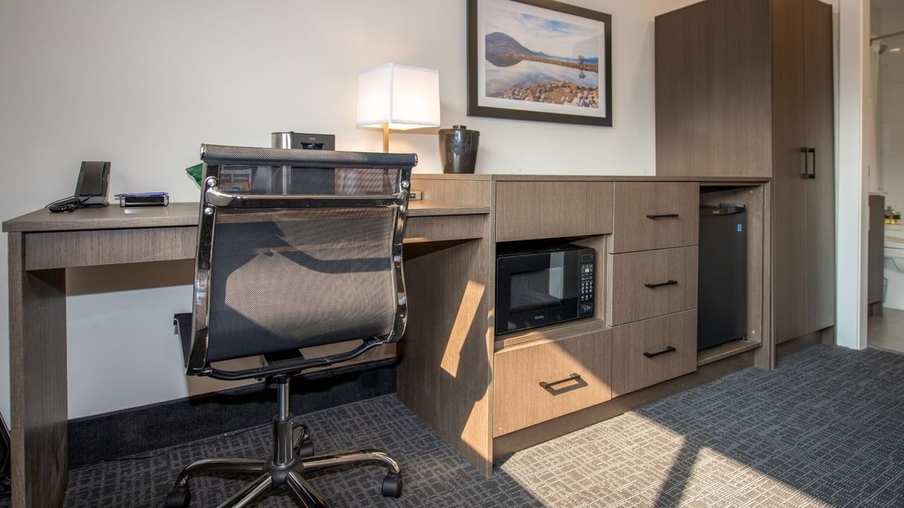 Hotel room with desk, chair, lamp, microwave, fridge, and picture frame on wall.