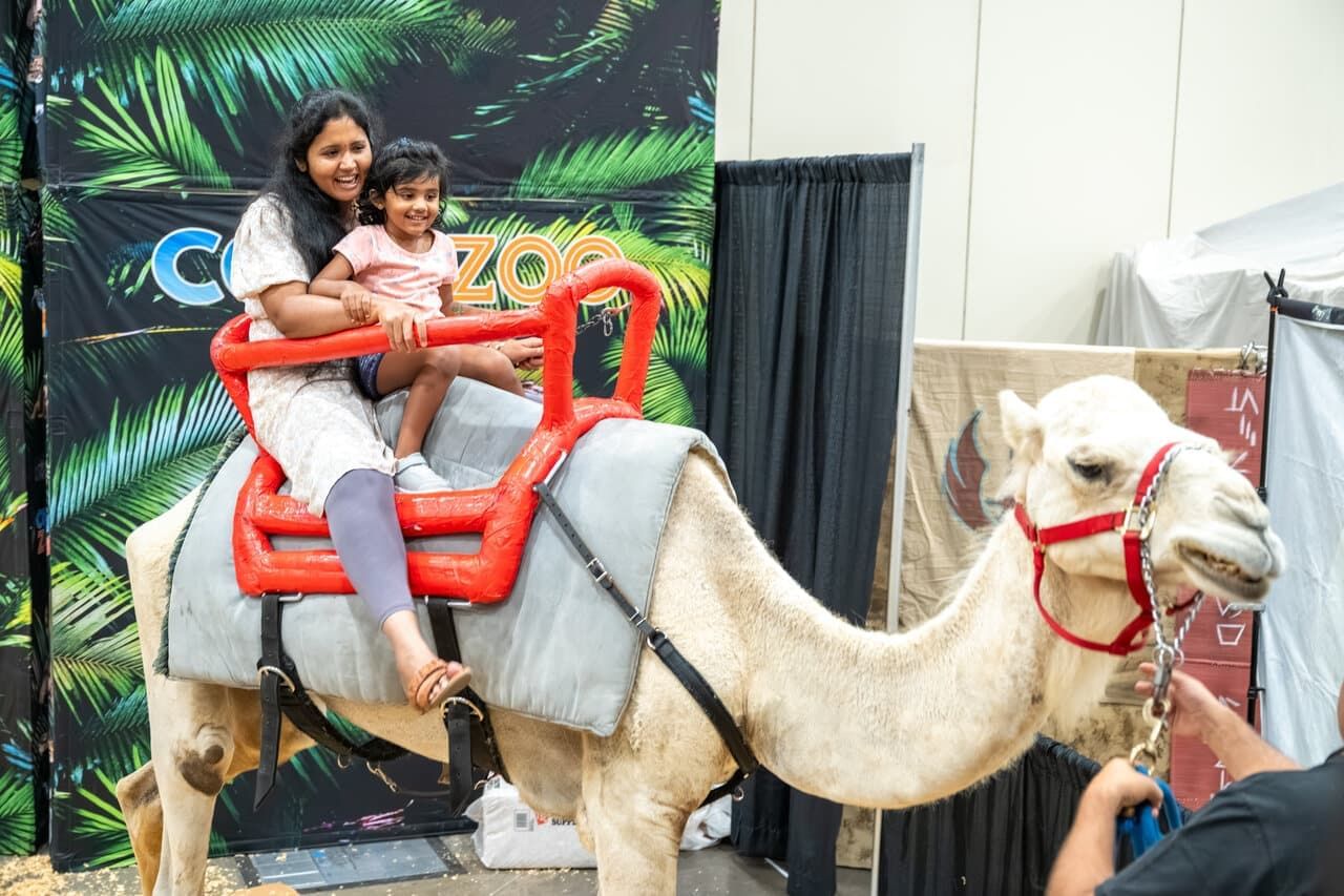 A woman holds a child while riding on the back of a camel in front of a tropical greenery sign.