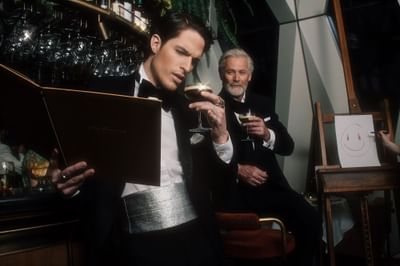 Man in tuxedo reads a menu while another guest raises a cocktail in a stylish bar with a smiley face at Hotel Motto Vienna