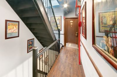Chelsea Pines NYC Hotel hallway with stairs, wooden floor, framed posters, brown door, and red pipe along the wall.