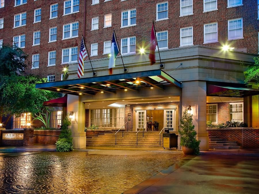 Exterior view of Warwick Melrose Dallas Hotel entrance at night with brick facade, flags, and covered driveway canopy