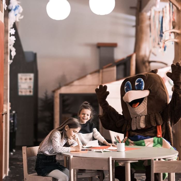 Zwei Mädchen und ein Maskottchen sitzen an einem Tisch mit Stiften und Papier.