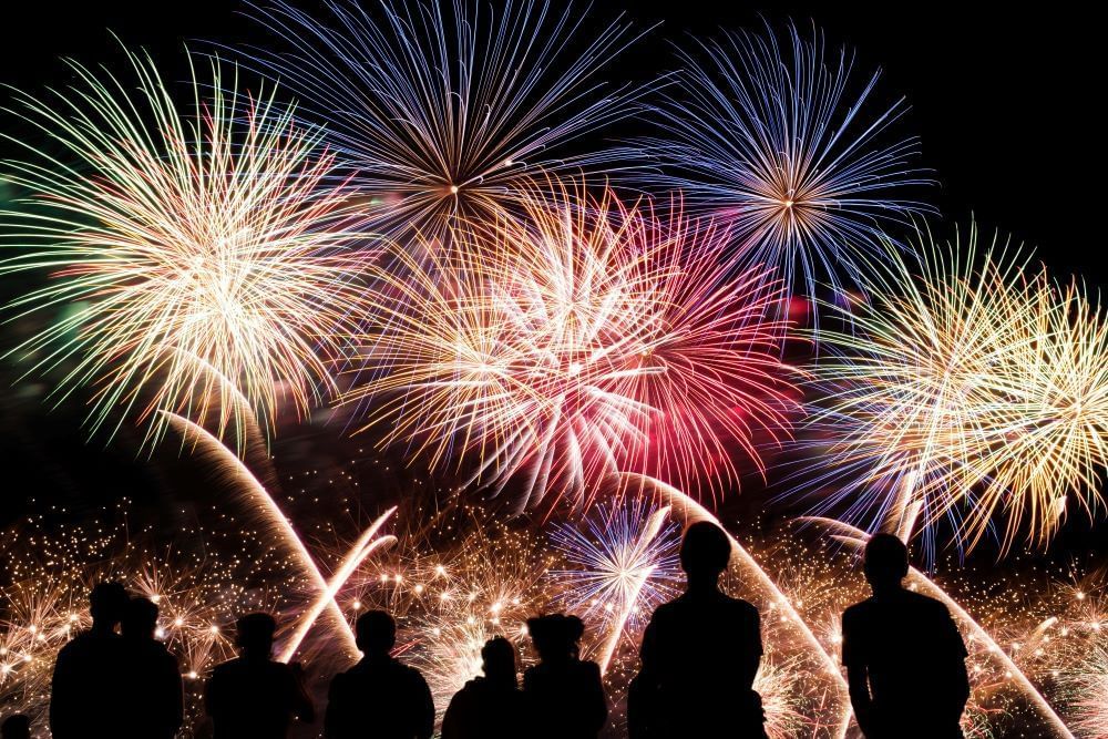 Silhouettes of people watch fireworks of white, gold, red, and blue burst in the black night sky.