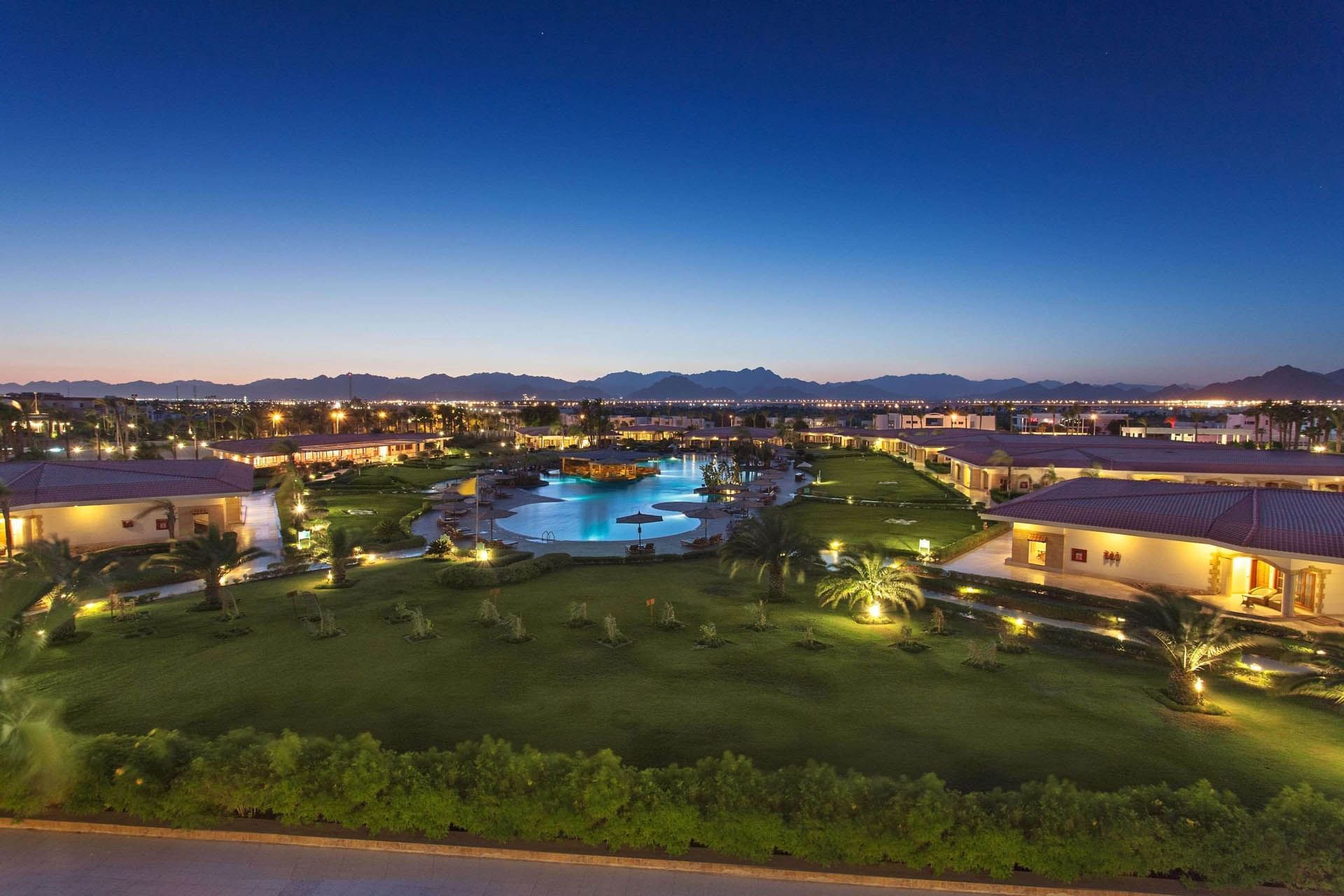 Serene dusk view of Pickalbatros Golf Beach Resort, with lush lawns, a glowing pool, palm trees, and distant mountains