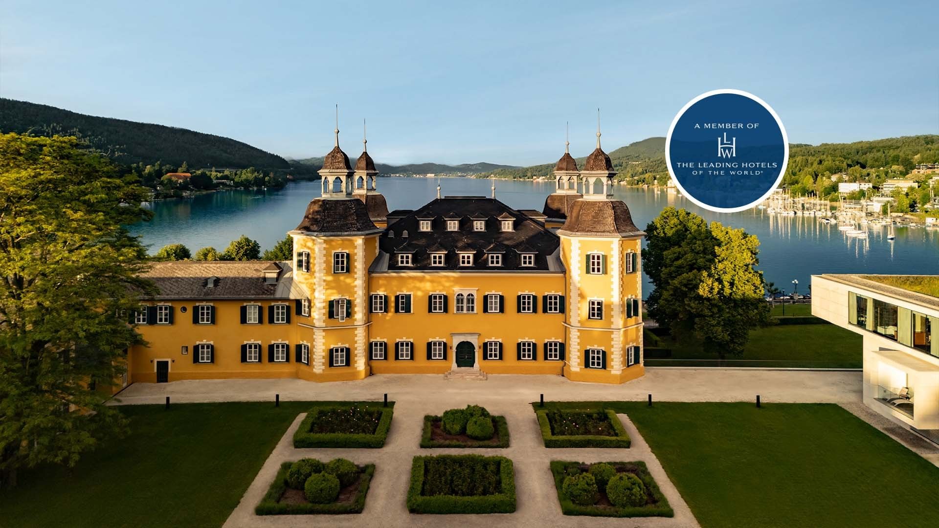 Gelbes Schloss mit Seeblick, umgeben von Gärten und einem modernen Gebäude.