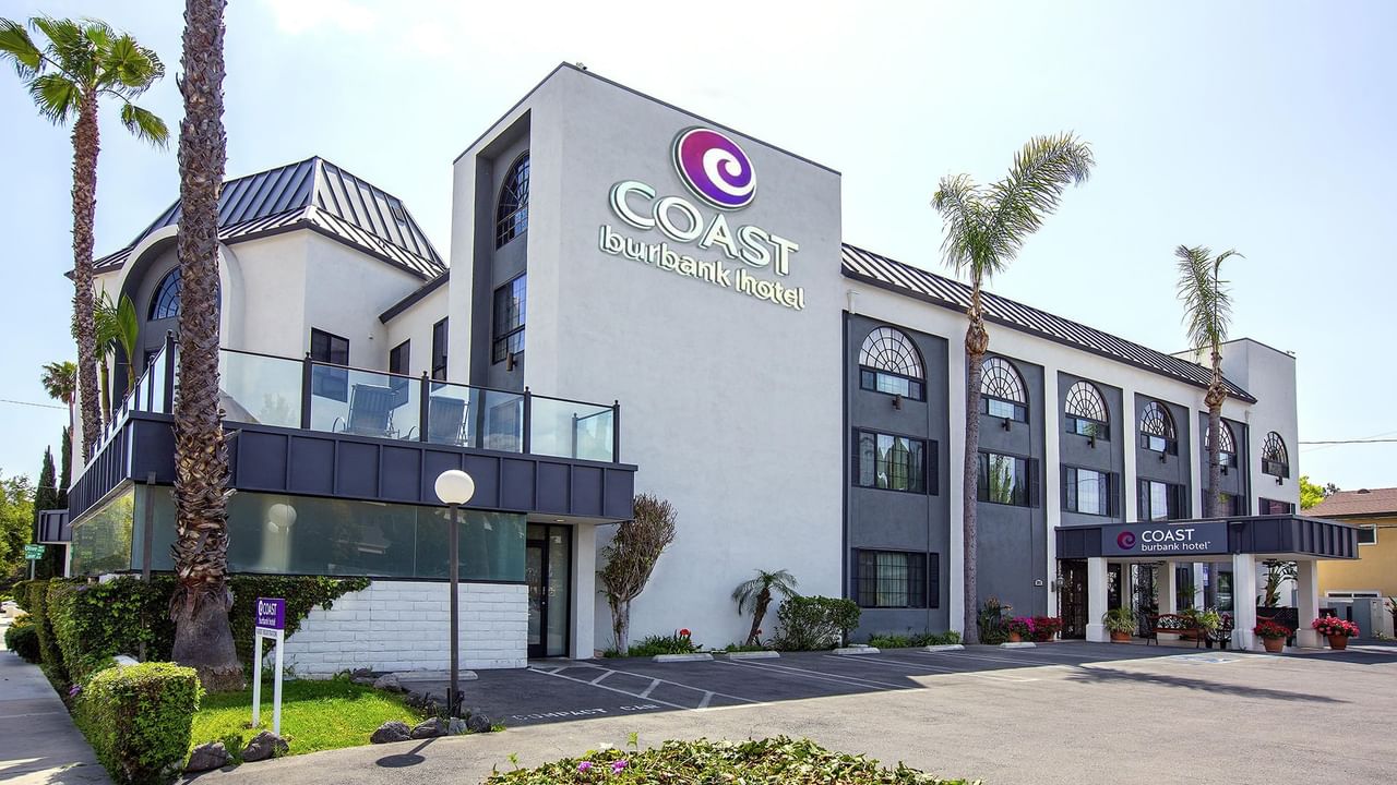 Exterior of Coast Burbank Hotel with palm trees, bushes, and a parking area.