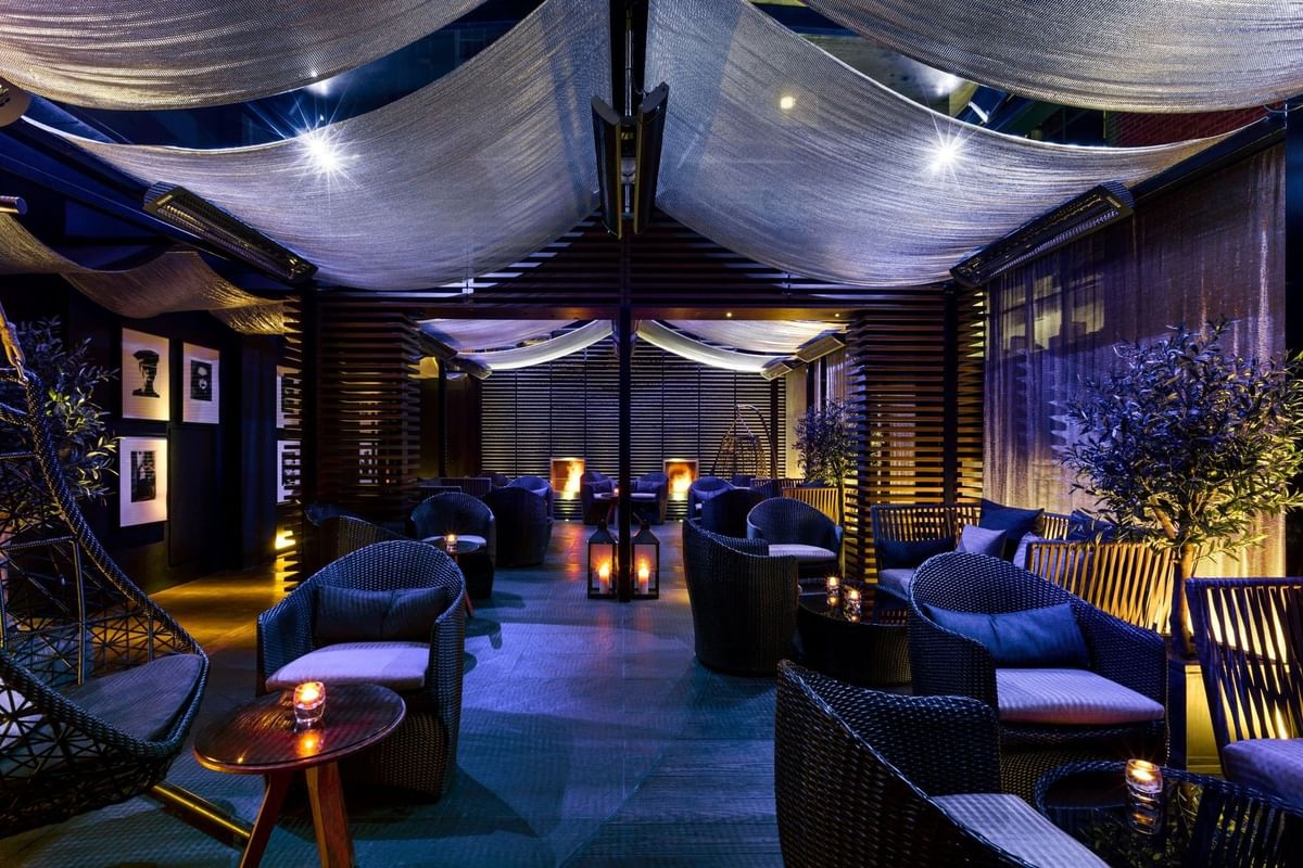 Blue-lit lounge with wicker chairs, hanging canopy, and warm candles in the May Fair Terrace at The May Fair Hotel.