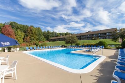 Fox Ridge Resort outdoor pool surrounded by blue lounge chairs, white tables, and a building on a hill under a blue sky.
