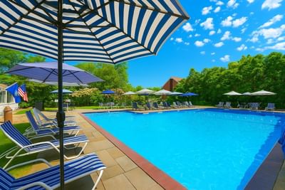 Outdoor blue swimming pool with striped loungers and umbrellas beside lush trees under clear sky at Avon Old Farms Hotel