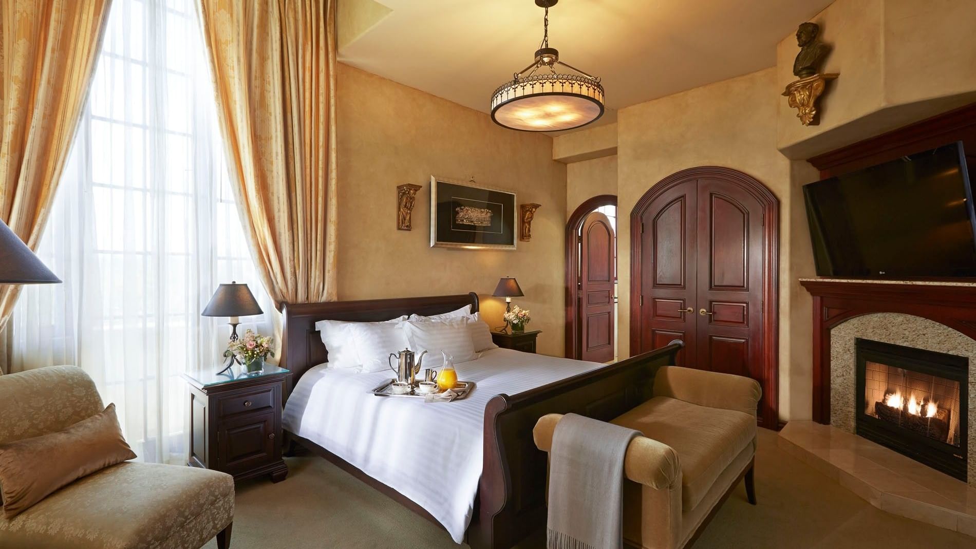 Sleigh bed by a fireplace under a round chandelier, featuring a tan bench in Kelly & Duane at The Mission Inn