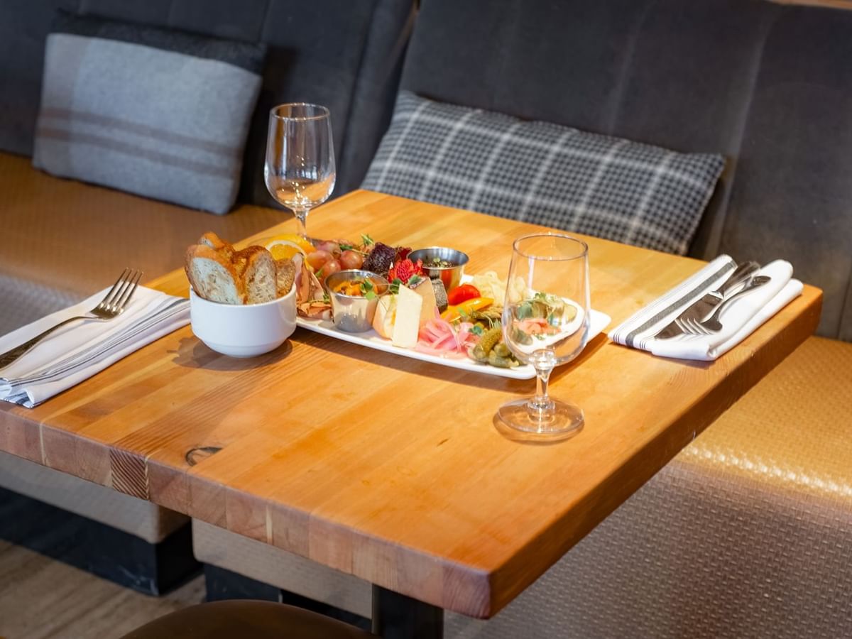 A wooden table set with a plate of food, bread, wine glasses, and silverware, adjacent to a cushioned bench.