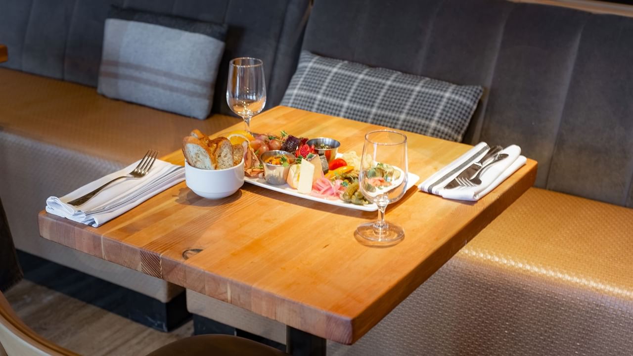 A wooden table set with a plate of food, bread, wine glasses, and silverware, adjacent to a cushioned bench.