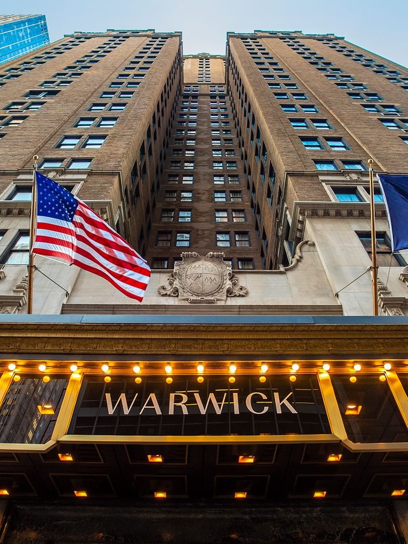 Upward view of historic hotel facade with illuminated marquee displaying Warwick name, flanked by American and hotel flags.