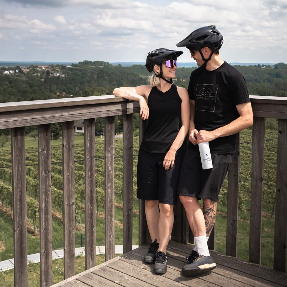Two cyclists in helmets pause on a scenic overlook with rolling hills and forests in the background.