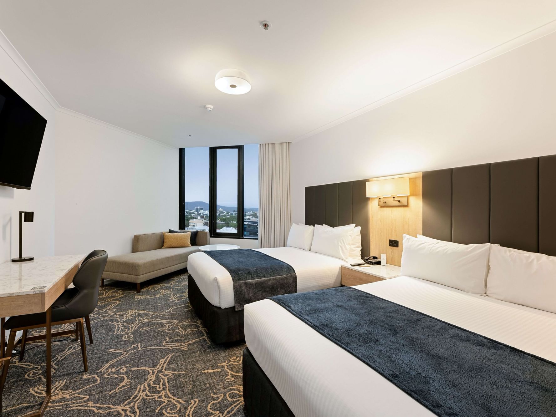 Twin room at Hotel Grand Chancellor Brisbane featuring two double beds, a marble-top work desk, wall-mounted TV, and a window overlooking the Brisbane city skyline.