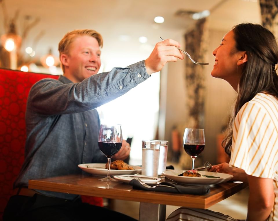 A man feeds a woman at Cora's Restaurant during a romantic dinner with wine and smiles at The White House Hotel