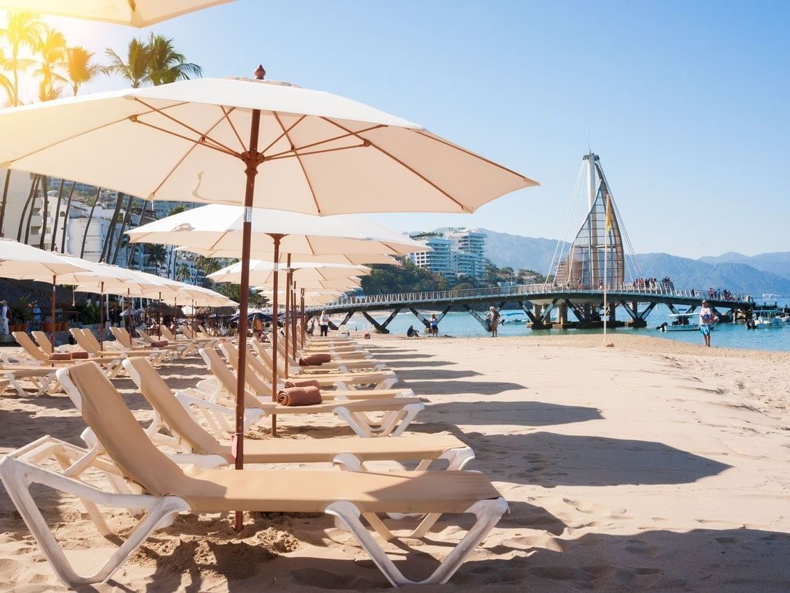 Rows of lounge chairs under umbrellas on a sandy shore facing the Los Muertos pier at Playa Los Arcos Beach Resort & Spa