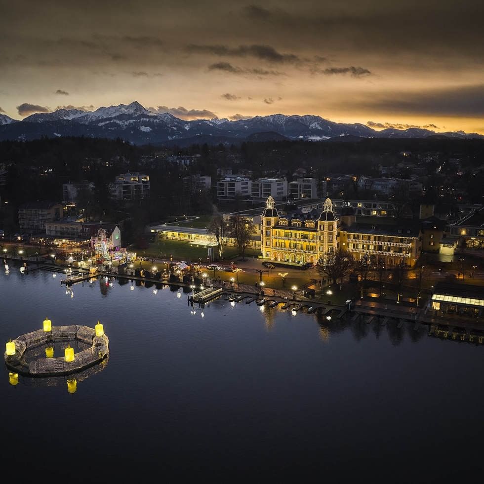 Illuminated yellow castle by lakeside with floating advent wreath and Christmas market at dusk with mountains beyond.