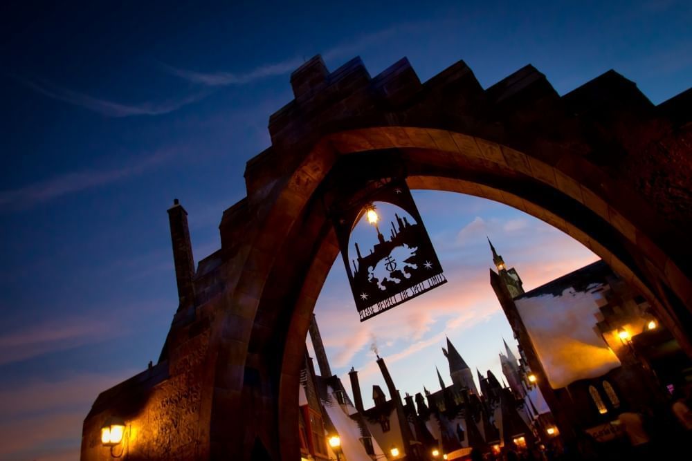 The arched entryway to Hogsmeade Village lit by glowing lamps and silhouetted against a twilight sky. 