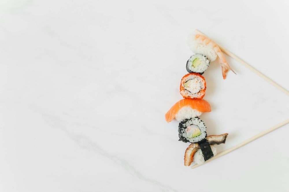 A simple white marble backdrop with crossed chopsticks holding a stack of different kinds of sushi. 