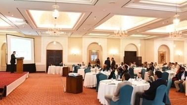 Speaker at a podium addressing guests at round tables in a large carpeted conference hall at Serena Hotels & Resorts