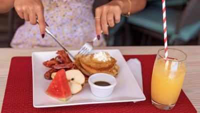 Person cutting pancakes with a fork and knife beside fresh watermelon and orange juice glass at Worthing Court Apartment