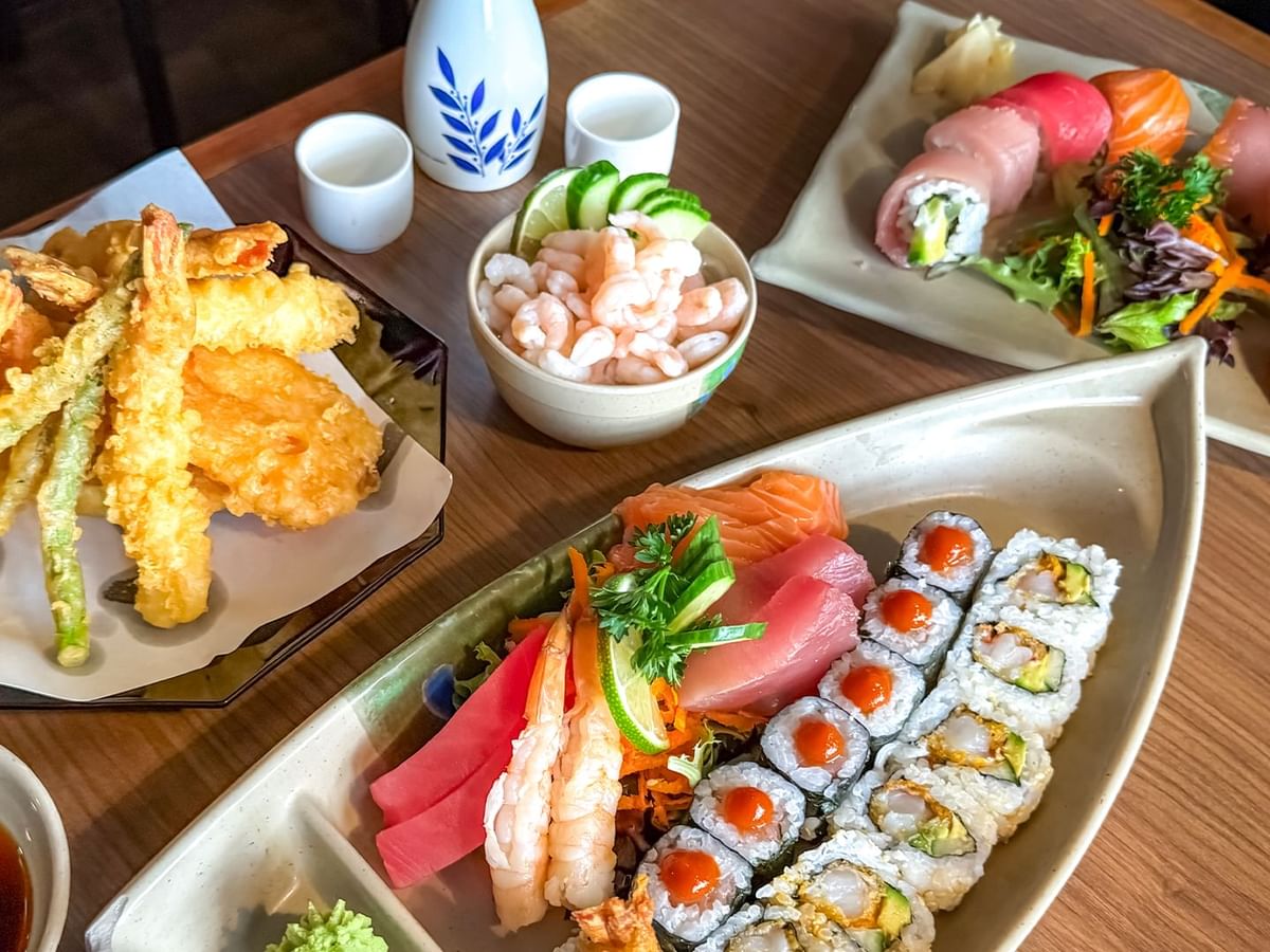 Plates of sushi, sashimi, tempura, and shrimp with dipping sauce on a wooden table.