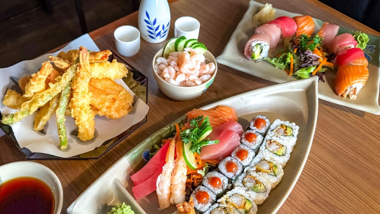 Plates of sushi, sashimi, tempura, and shrimp with dipping sauce on a wooden table.