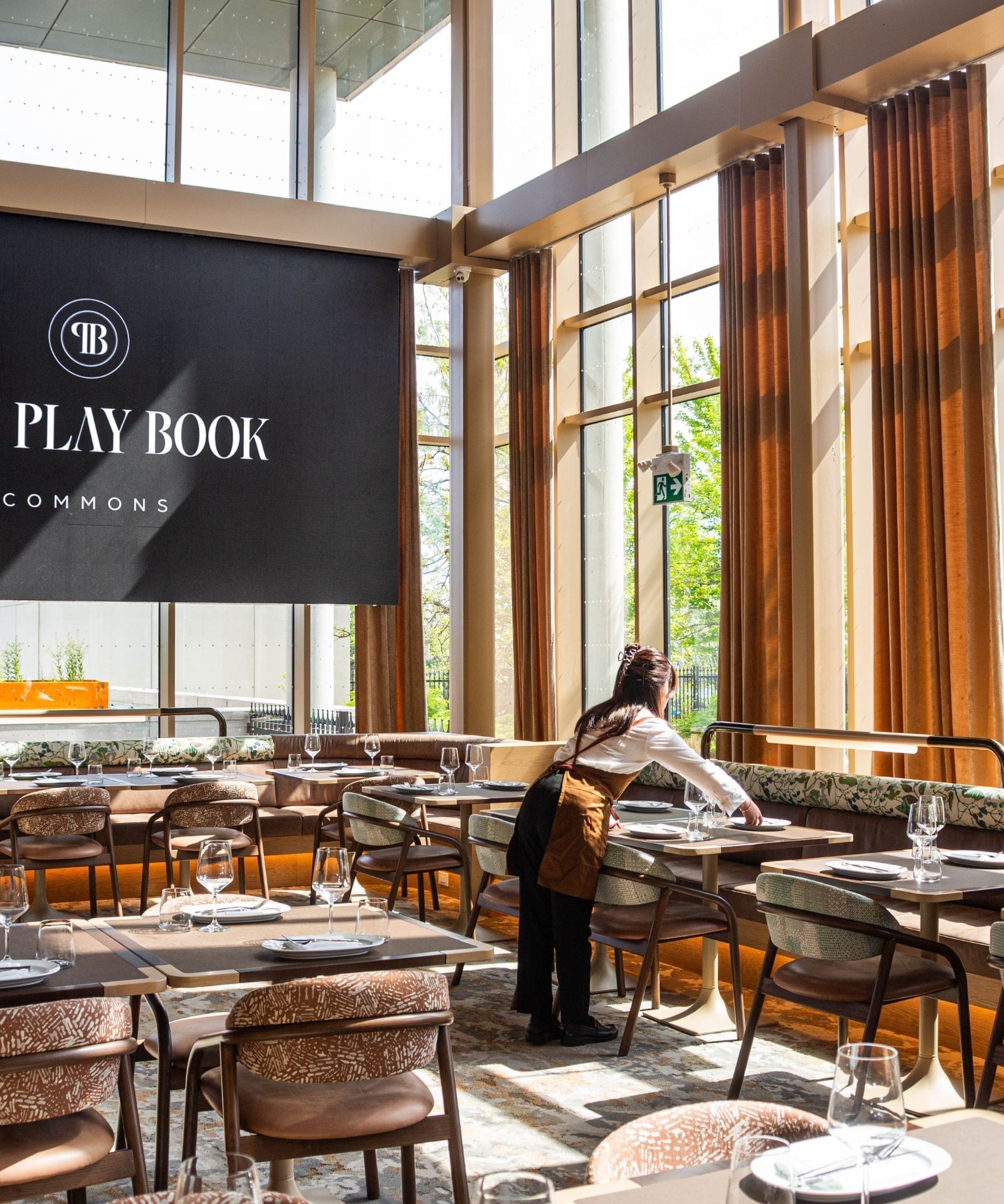 Play Book Commons dining room with set tables and patterned chairs beside tall sunlit windows at Hotel X Toronto