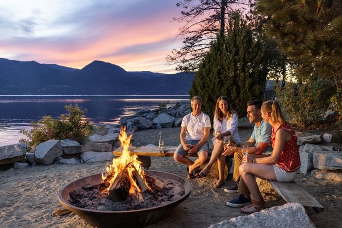 Group of four friends sitting around a campfire at sunset near a lake, enjoying drinks and conversation.