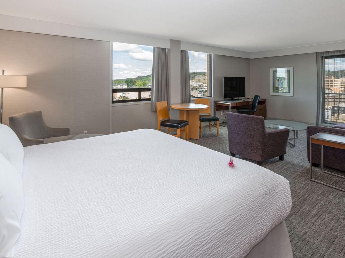 A hotel suite with a king bed, seating area, desk, and balcony with city view.